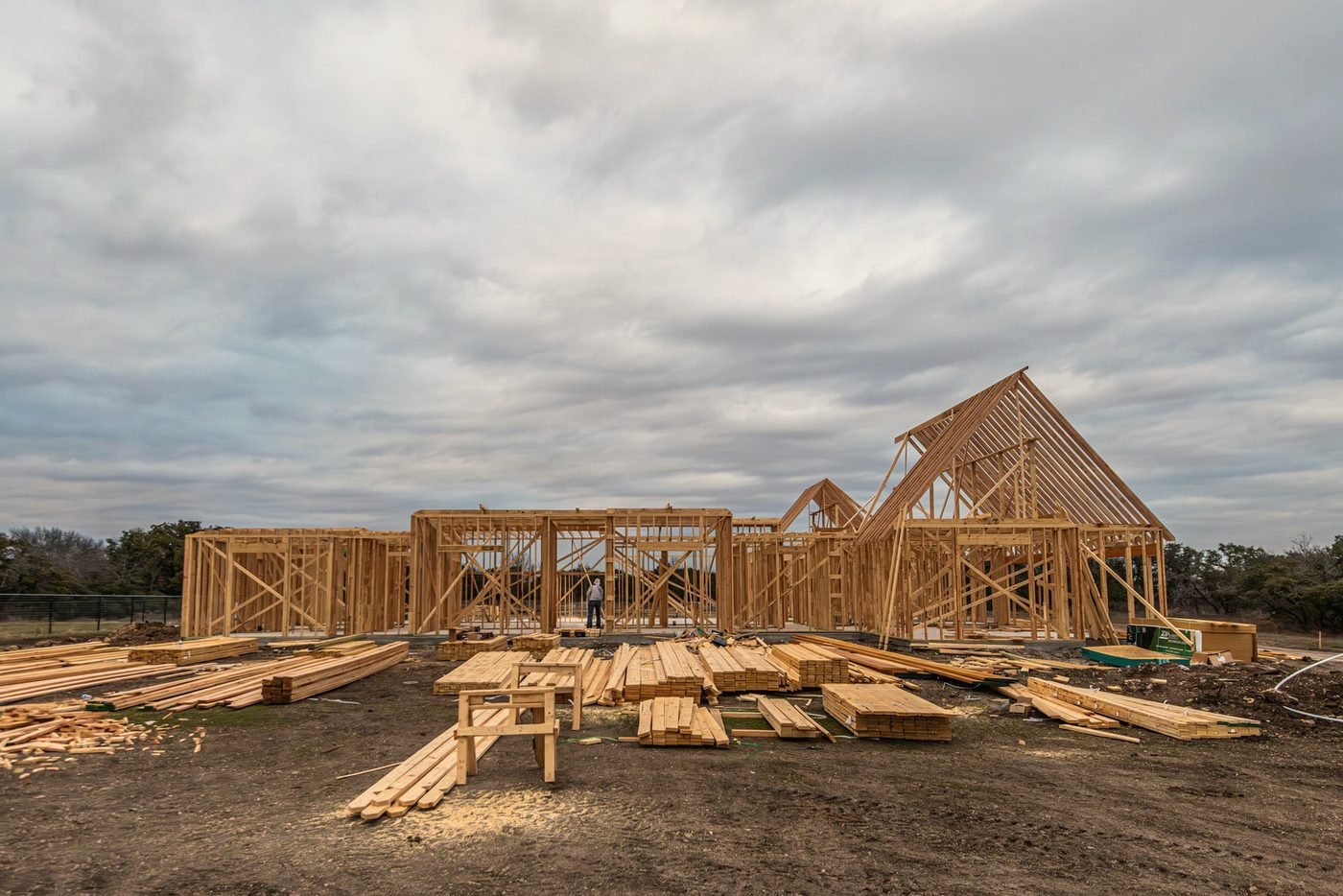 A house frame going up on open ground under a grey plains sky with lumber stacked on site.